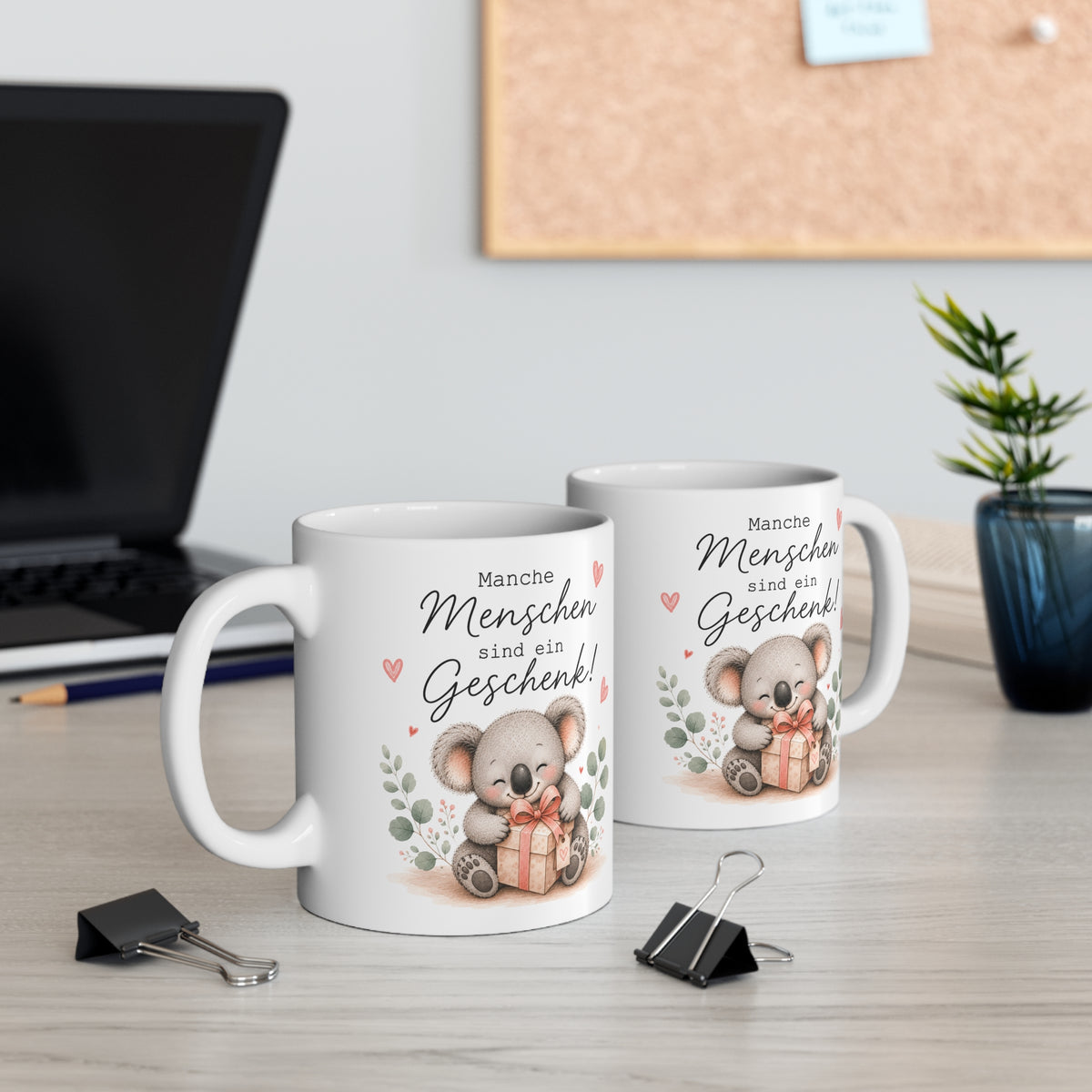 Zwei weiße Keramiktassen mit süßem Koala und Geschenkmotiv sowie dem Spruch „Manche Menschen sind ein Geschenk!“ auf einem Schreibtisch neben Laptop und Pflanze