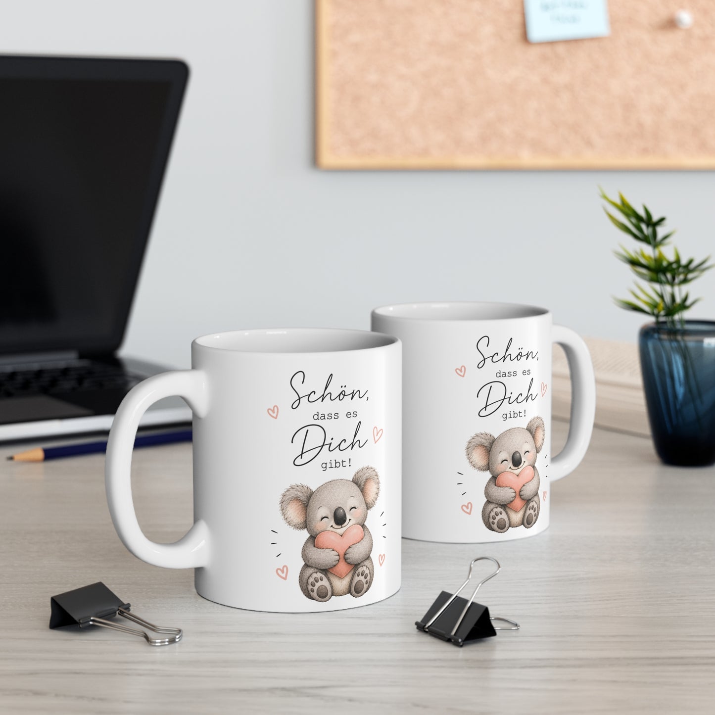 Zwei weiße Keramiktassen mit süßem Koala, Herzmotiv und dem Spruch „Schön, dass es Dich gibt!“ auf einem Schreibtisch neben Laptop und Pflanze