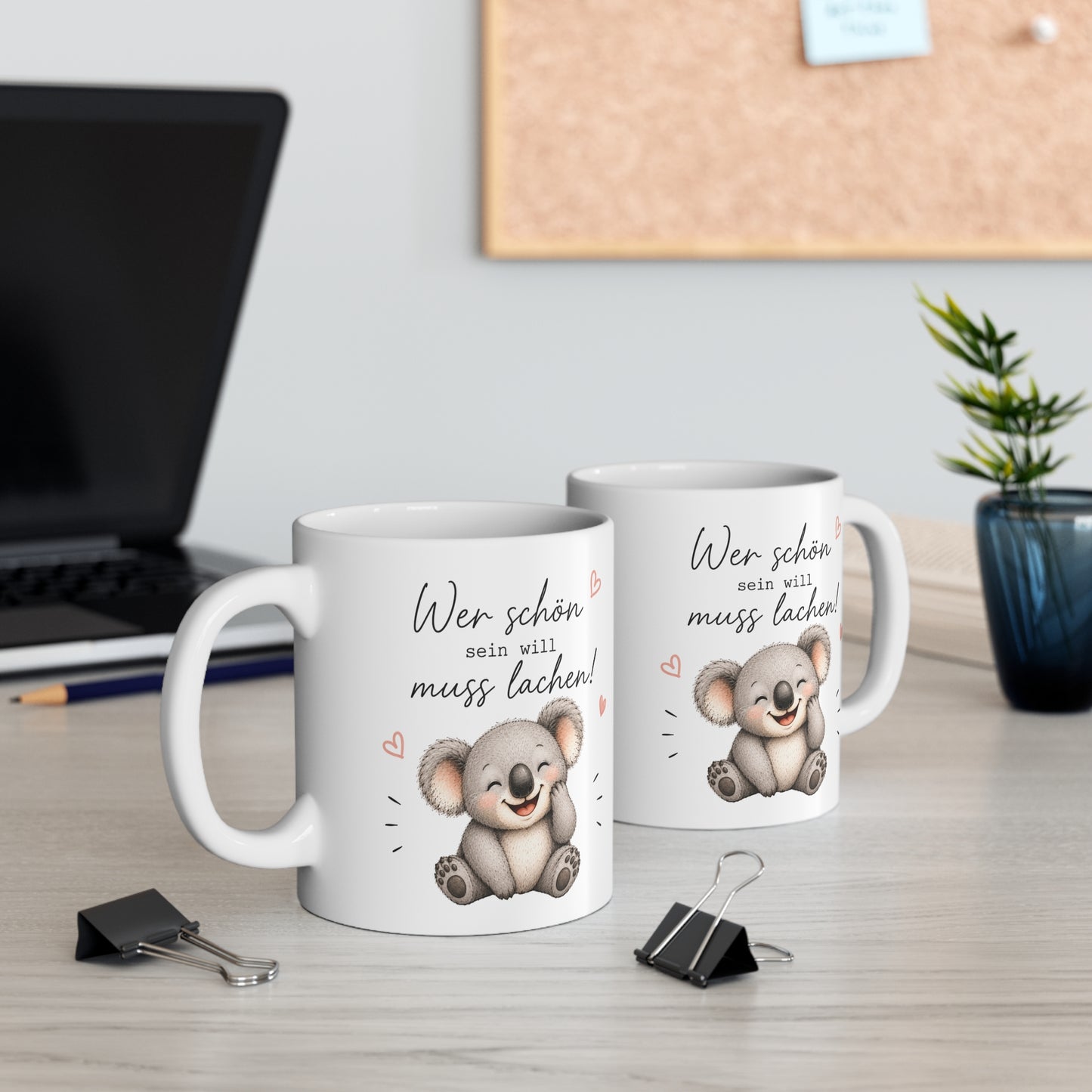Zwei weiße Keramiktassen mit lachendem Koala und dem Spruch „Wer schön sein will muss lachen“ auf einem Schreibtisch neben Laptop und Pflanze