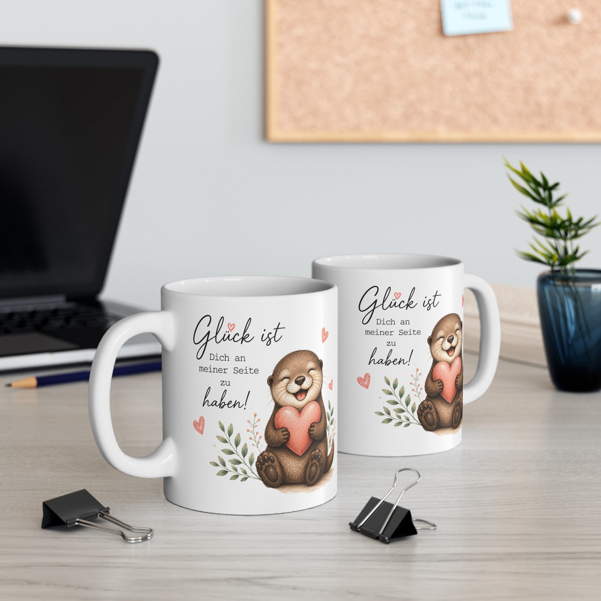 Zwei weiße Keramiktassen mit lächelndem Otter, Herzmotiv und dem Spruch „Glück ist Dich an meiner Seite zu haben“ auf einem Schreibtisch neben Laptop und Pflanze