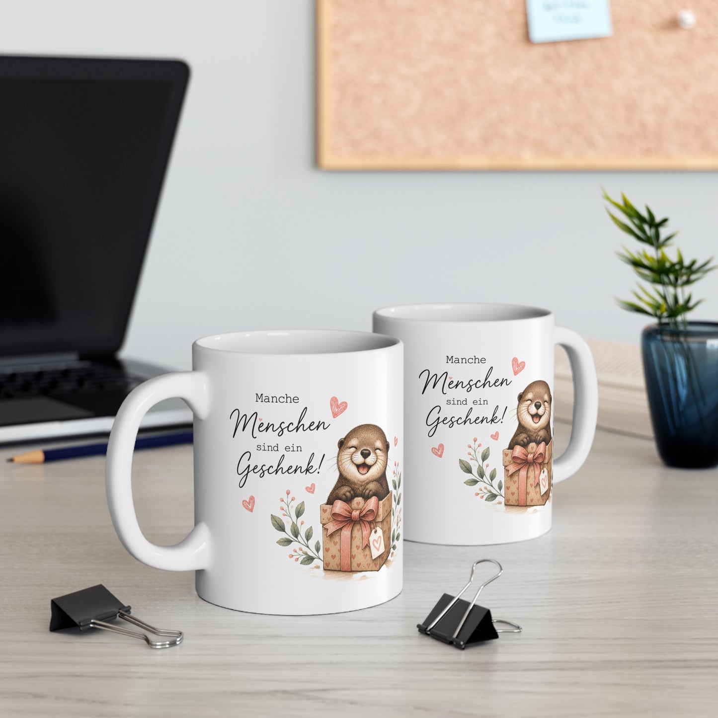 Zwei weiße Keramiktassen mit süßem Otter im Geschenkpaket und dem Spruch „Manche Menschen sind ein Geschenk“ auf einem Schreibtisch neben Laptop und Pflanze