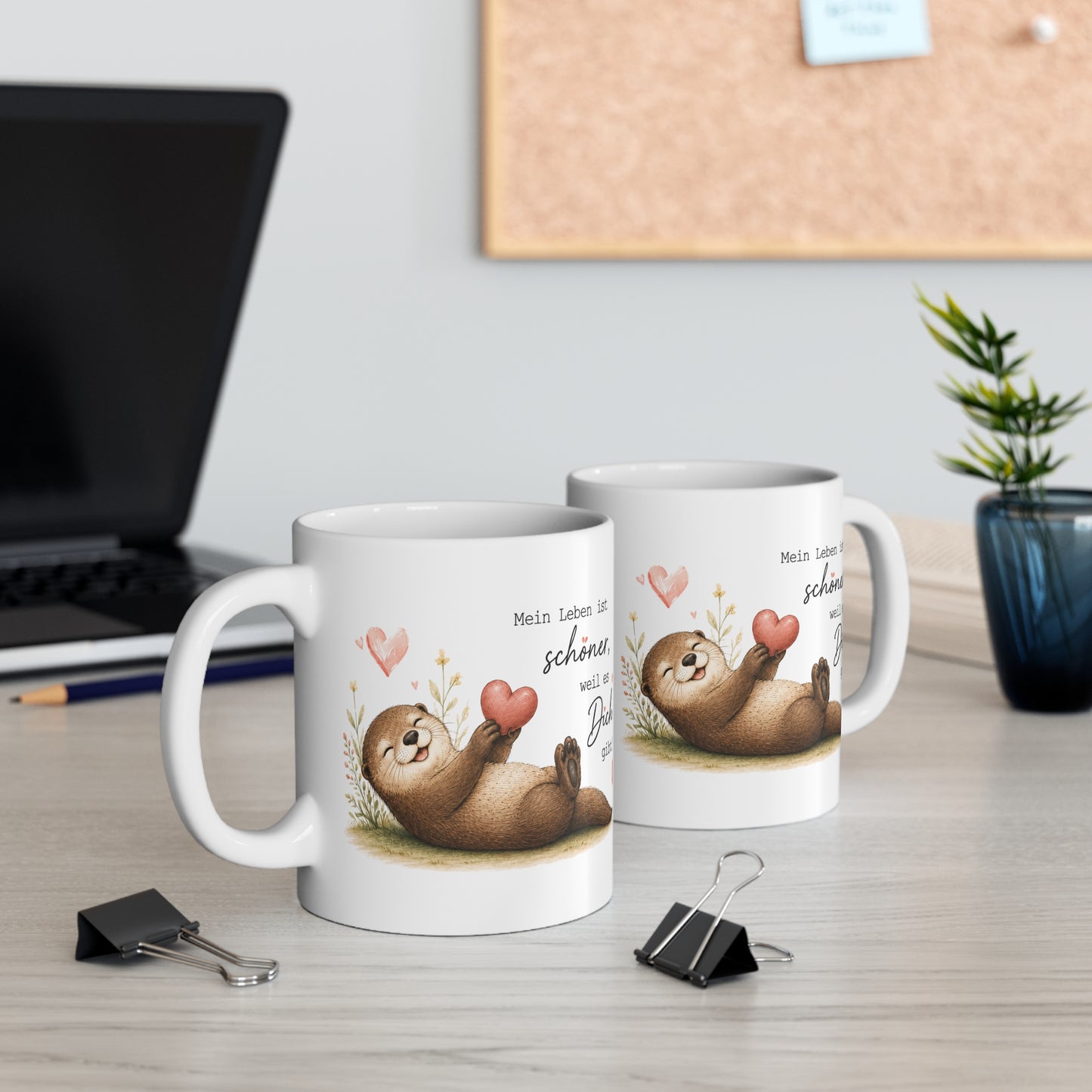 Zwei weiße Keramiktassen mit liegendem Otter, Herzmotiv und dem Spruch „Mein Leben ist schöner, weil es Dich gibt“ auf einem Schreibtisch
