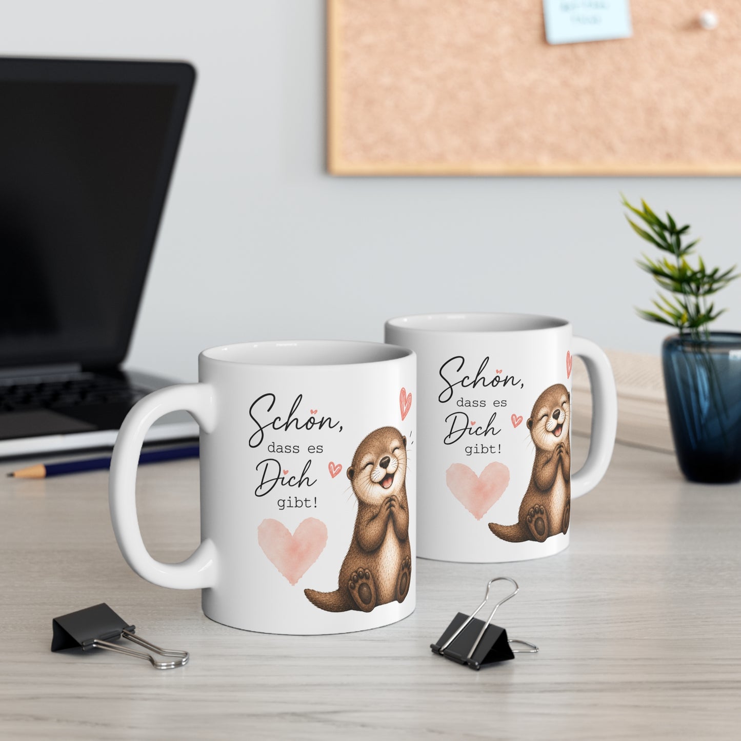 Zwei weiße Keramiktassen mit lächelndem Otter, Herzmotiven und dem Spruch „Schön, dass es Dich gibt“ auf einem Schreibtisch neben Laptop und Pflanze