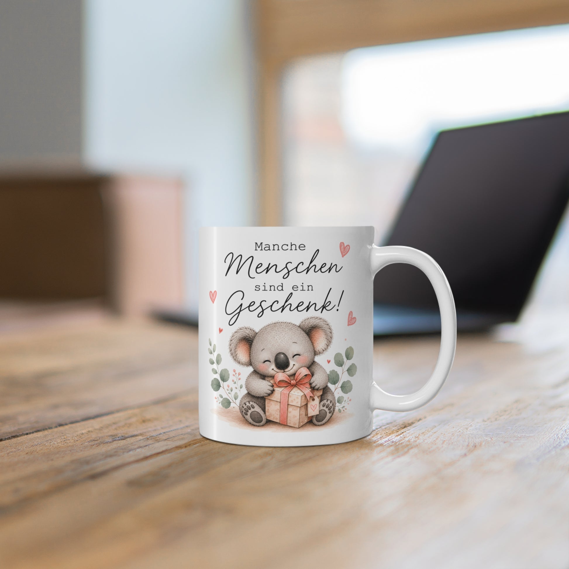 Weiße Tasse mit niedlichem Koala, Geschenkbox und dem Schriftzug „Manche Menschen sind ein Geschenk!“ auf einem Holztisch