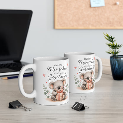 Zwei weiße Keramiktassen mit süßem Koala und Geschenkmotiv sowie dem Spruch „Manche Menschen sind ein Geschenk!“ auf einem Schreibtisch neben Laptop und Pflanze