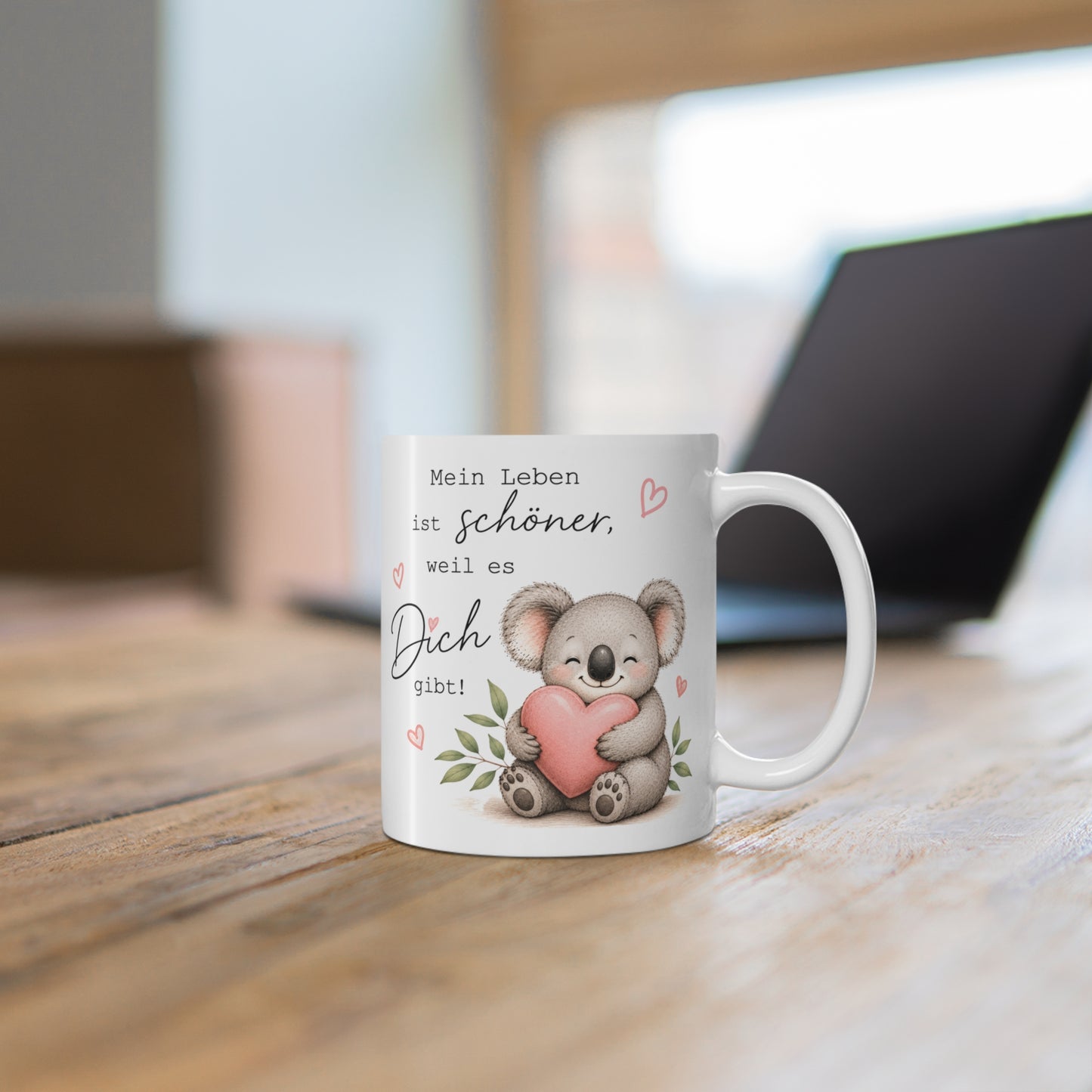 Weiße Tasse mit süßem Koala, rosa Herz und liebevollem Spruch auf einem Holztisch vor unscharfem Hintergrund