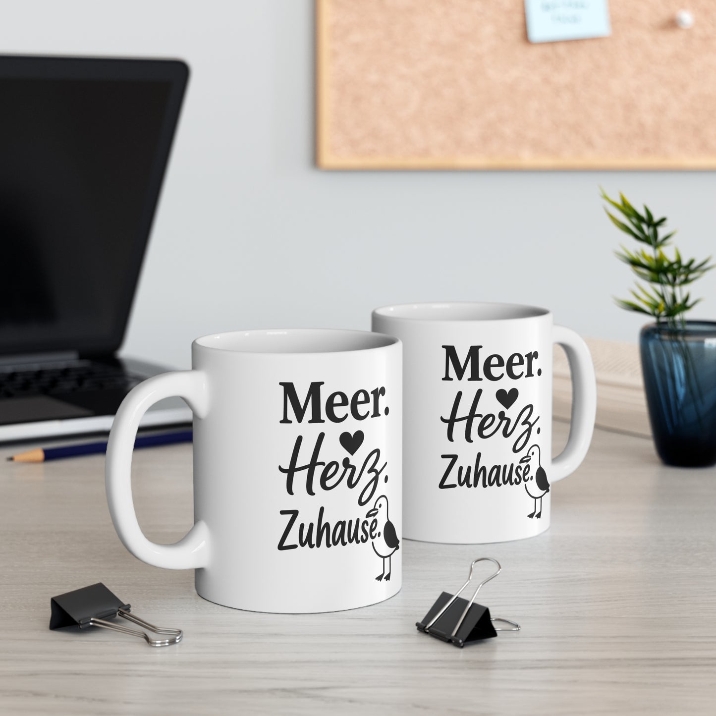 Zwei weiße Keramiktassen mit maritimem Spruch „Meer. Herz. Zuhause.“ und Möwenmotiv auf einem Schreibtisch