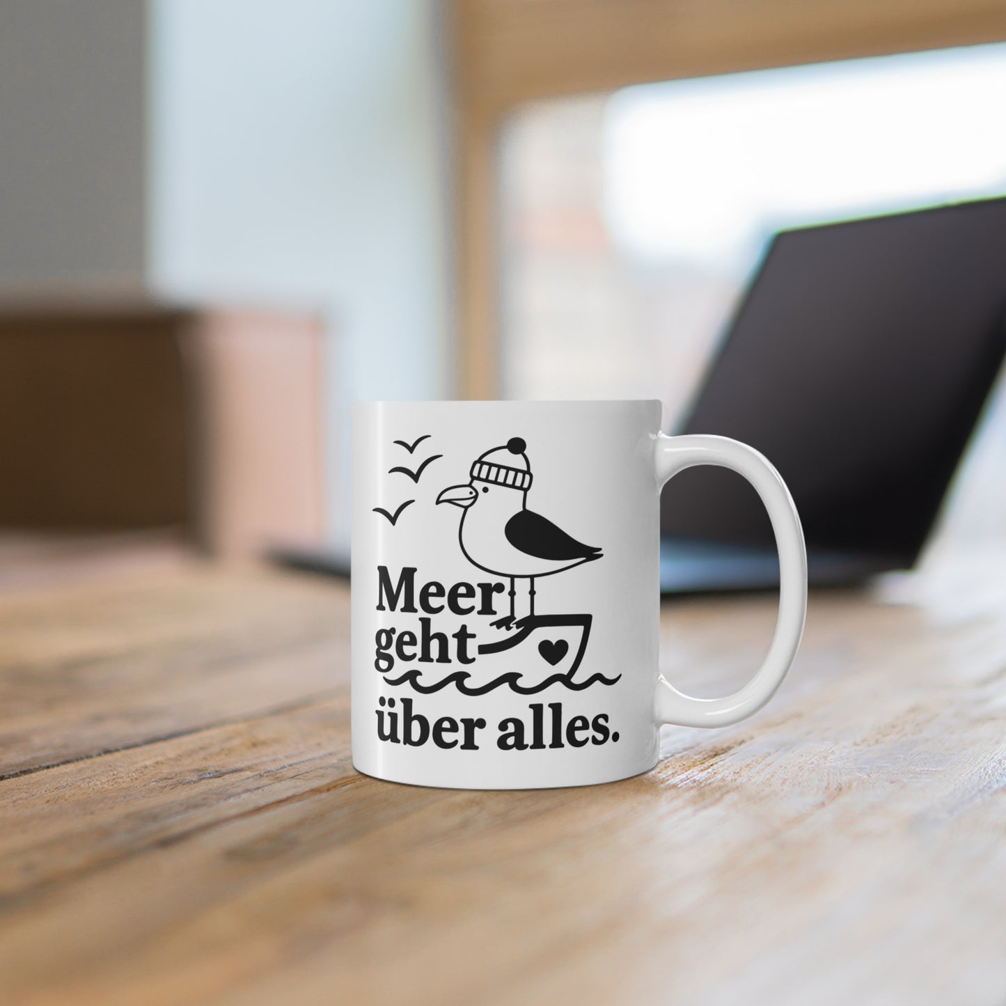 Weiße Tasse mit maritimer Möwe und Schriftzug „Meer geht über alles“ auf einem Holztisch vor unscharfem Hintergrund