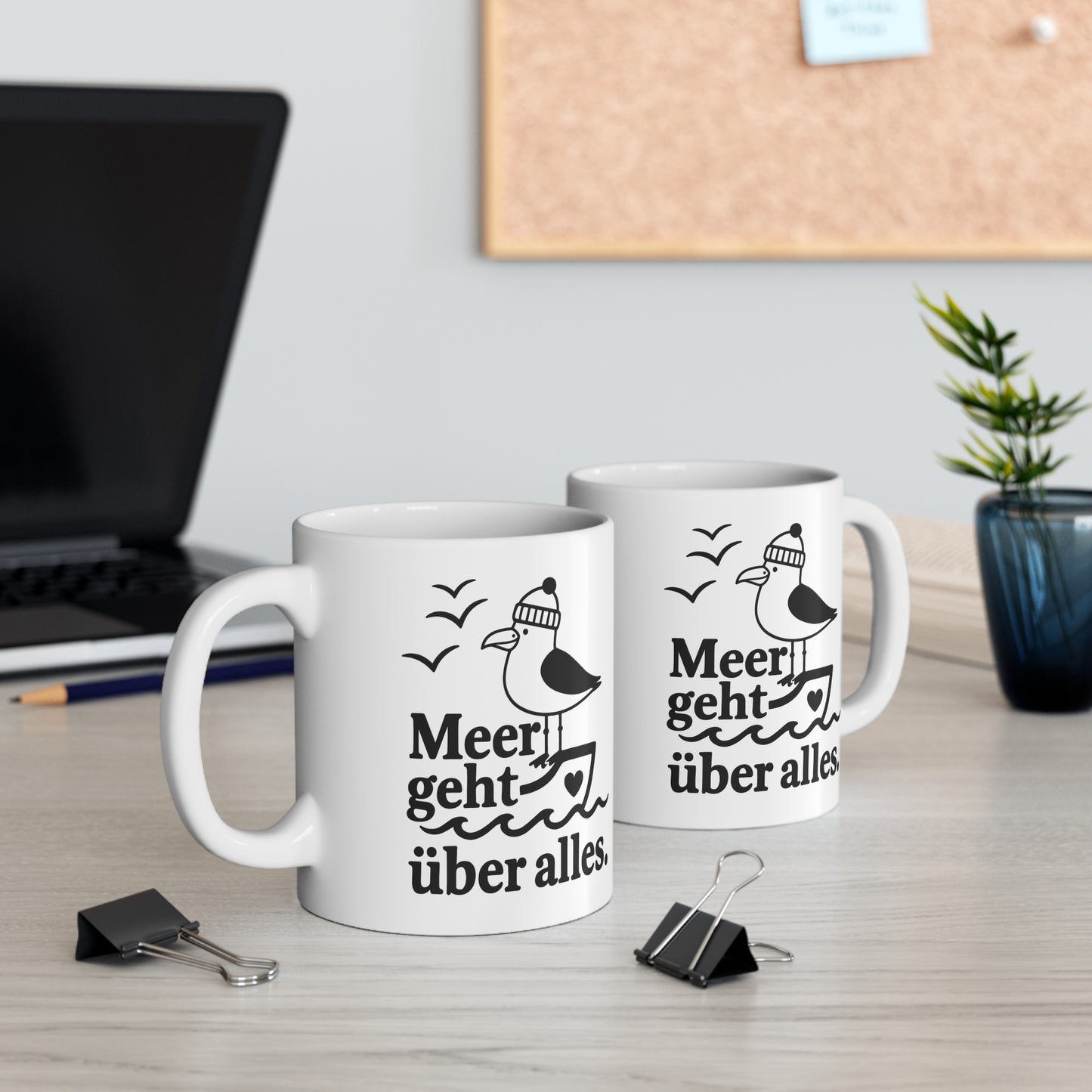 Zwei weiße Keramiktassen mit Möwenmotiv und Spruch „Meer geht über alles“ auf einem Schreibtisch neben Laptop und Pflanze