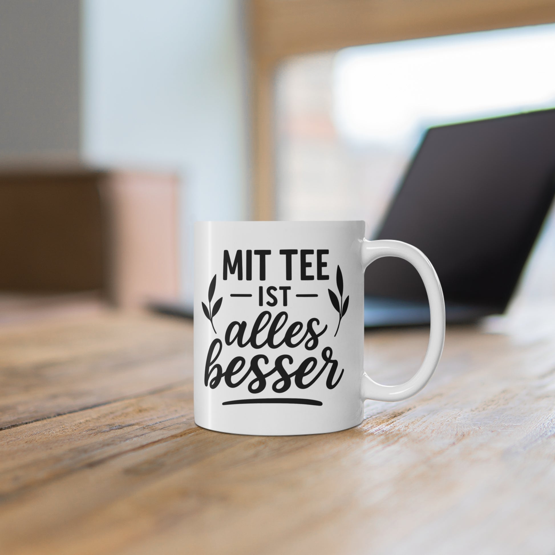 Weiße Keramiktasse mit Teebeutel-Motiv und Spruch „Mit Tee ist alles besser“ auf einem Schreibtisch neben Laptop und Pflanze