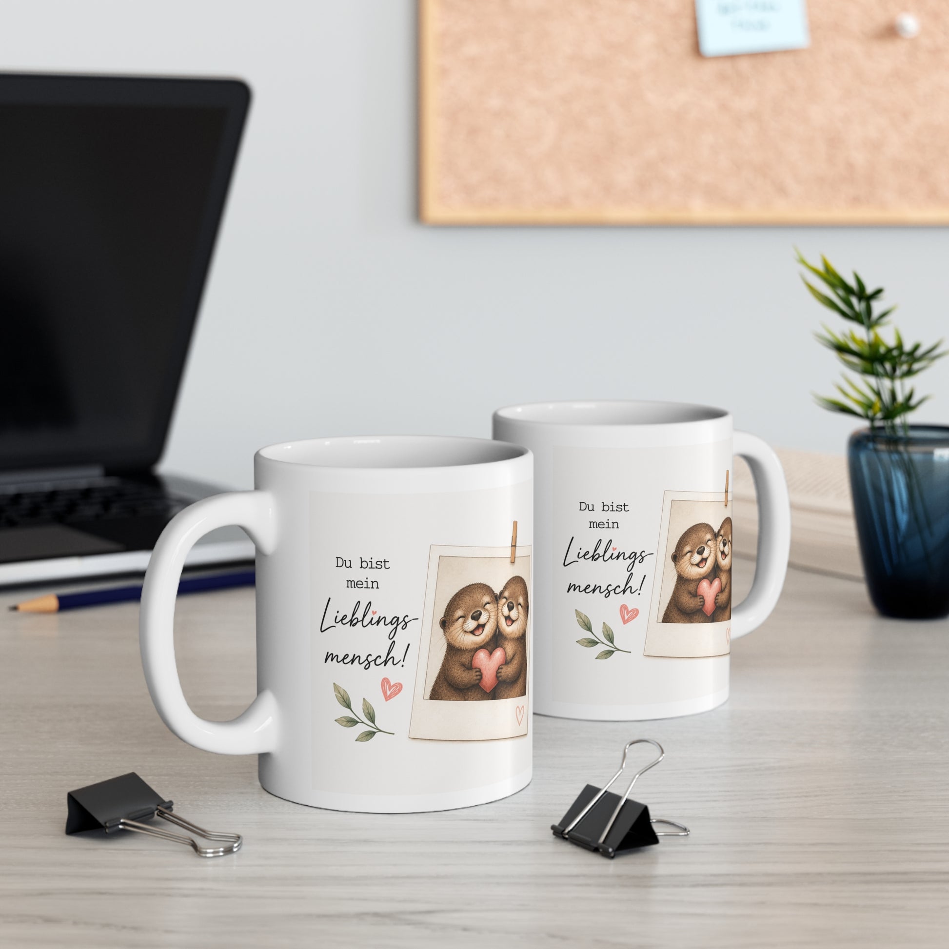 Zwei weiße Keramiktassen mit Otter-Pärchen, Herzmotiv und dem Spruch „Du bist mein Lieblingsmensch“ auf einem Schreibtisch neben Laptop und Pflanze