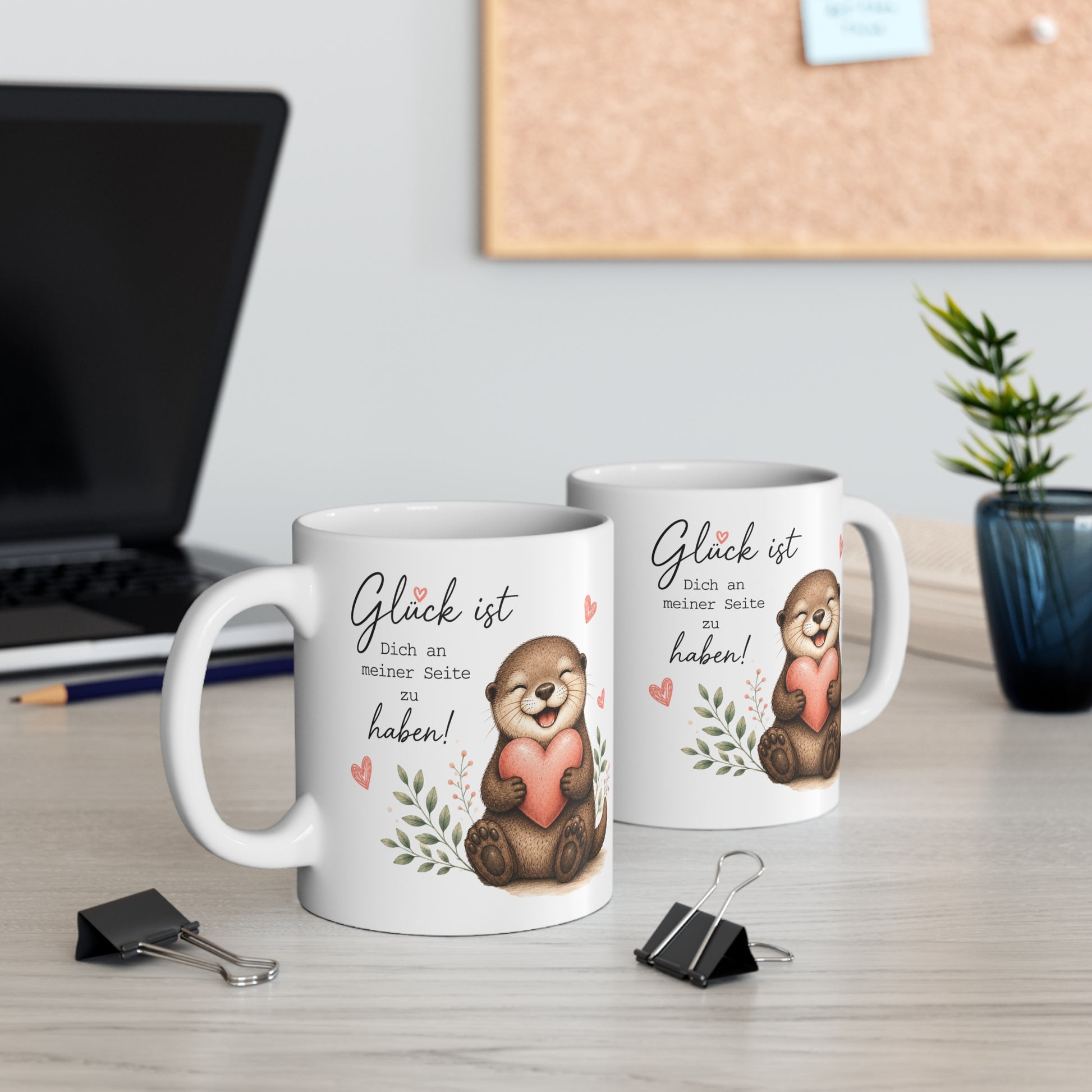 Zwei weiße Keramiktassen mit lächelndem Otter, Herzmotiv und dem Spruch „Glück ist Dich an meiner Seite zu haben“ auf einem Schreibtisch neben Laptop und Pflanze