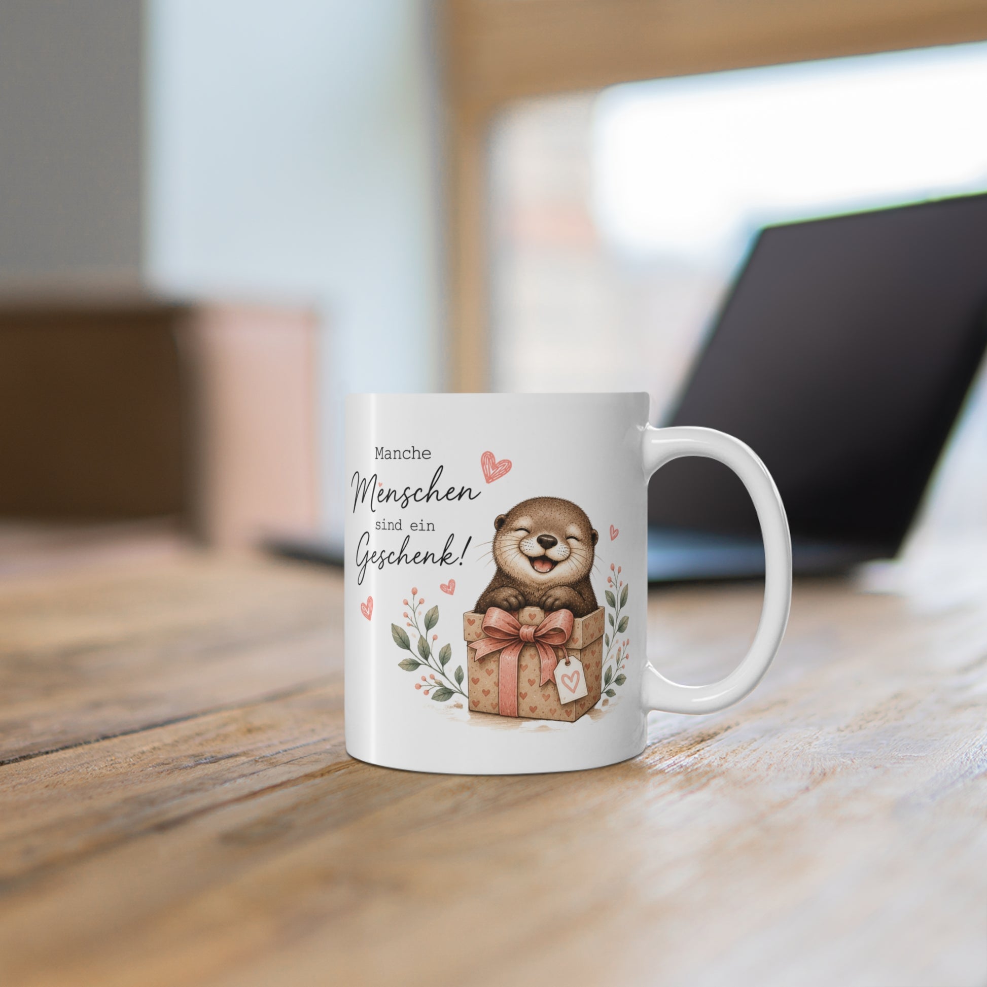 Weiße Tasse mit niedlichem Otter in einer Geschenkbox, Herzmotiven und dem Spruch „Manche Menschen sind ein Geschenk“ auf einem Holztisch