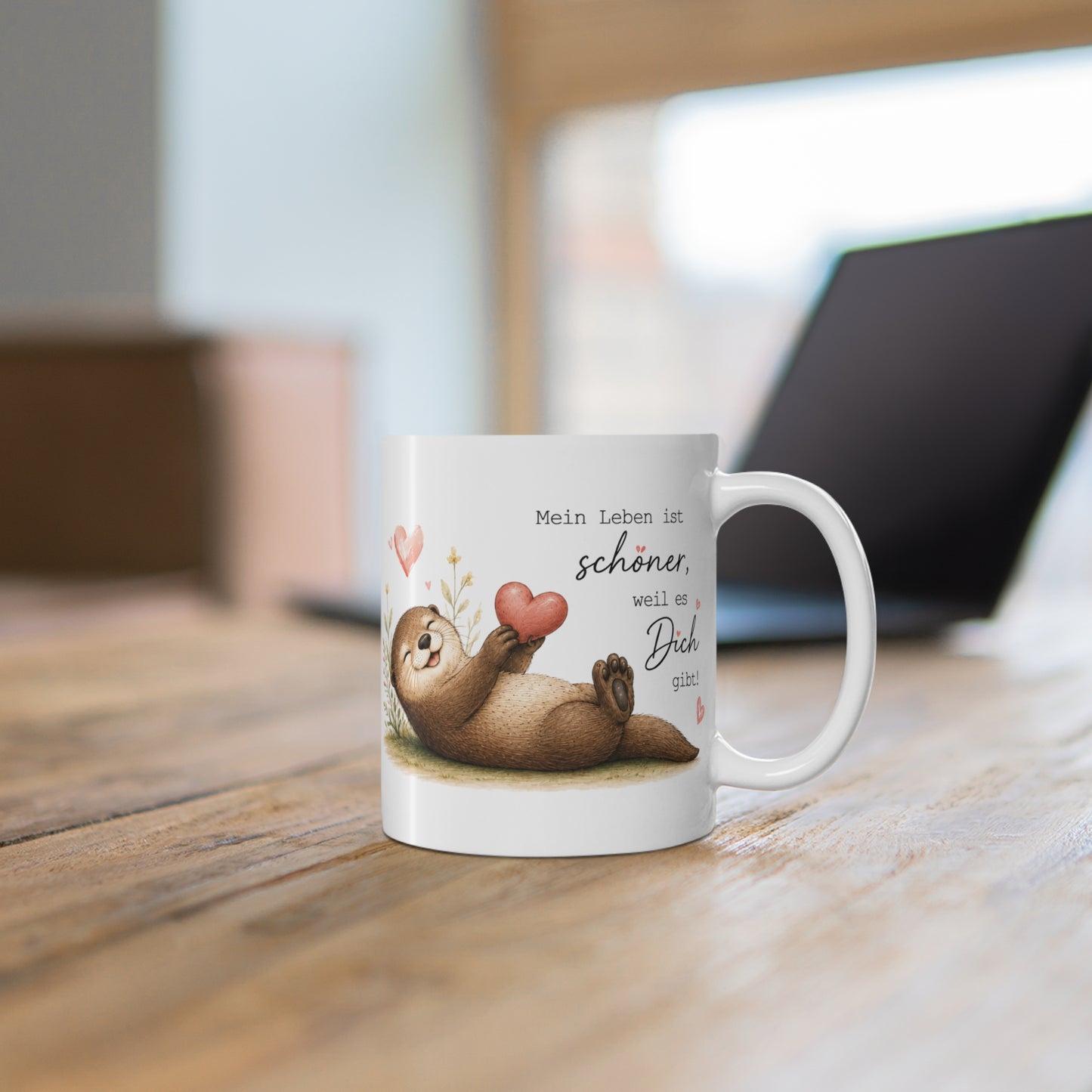 Weiße Tasse mit liegendem Otter, Herzillustration und liebevollem Spruch auf einem Holztisch vor verschwommenem Hintergrund