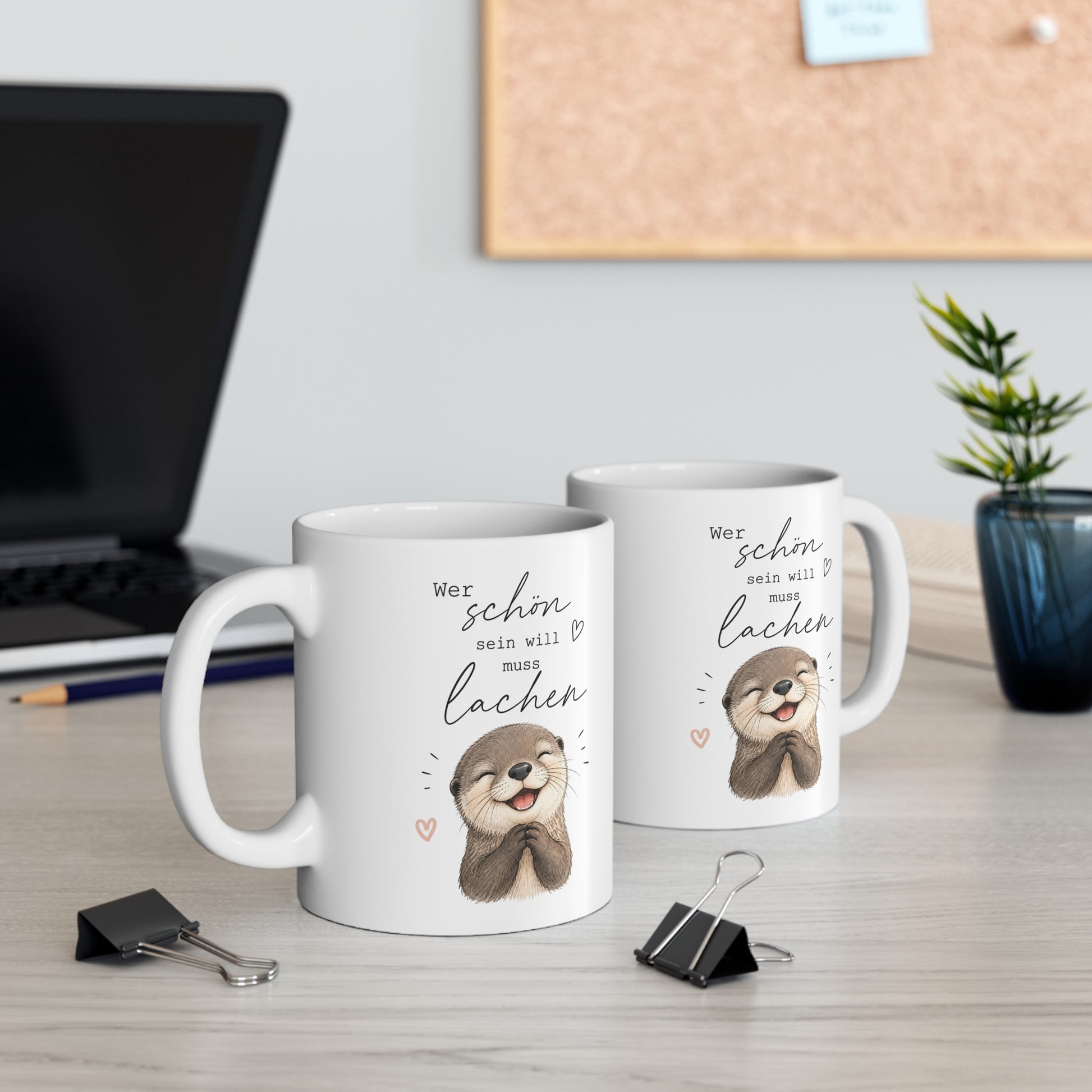 Zwei weiße Keramiktassen mit lachendem Otter und dem Spruch „Wer schön sein will muss lachen“ auf einem Schreibtisch neben Laptop und Pflanze