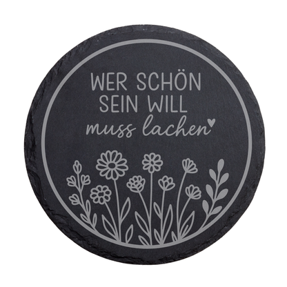 Runder schwarzer Schiefer-Untersetzer mit lustigem Spruch „Wer schön sein will muss lachen“ und floraler Illustration